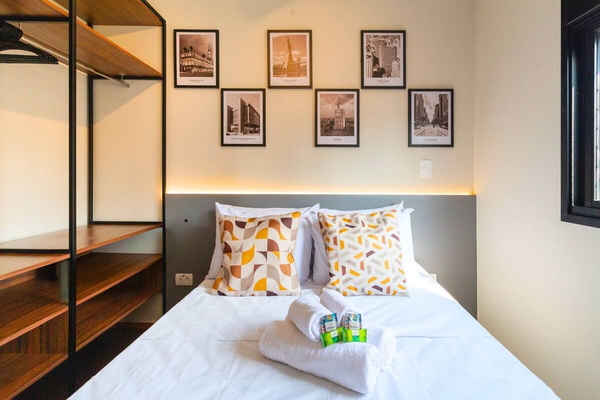 A hotel room with a bed, a wooden shelf, and a wall with framed pictures.