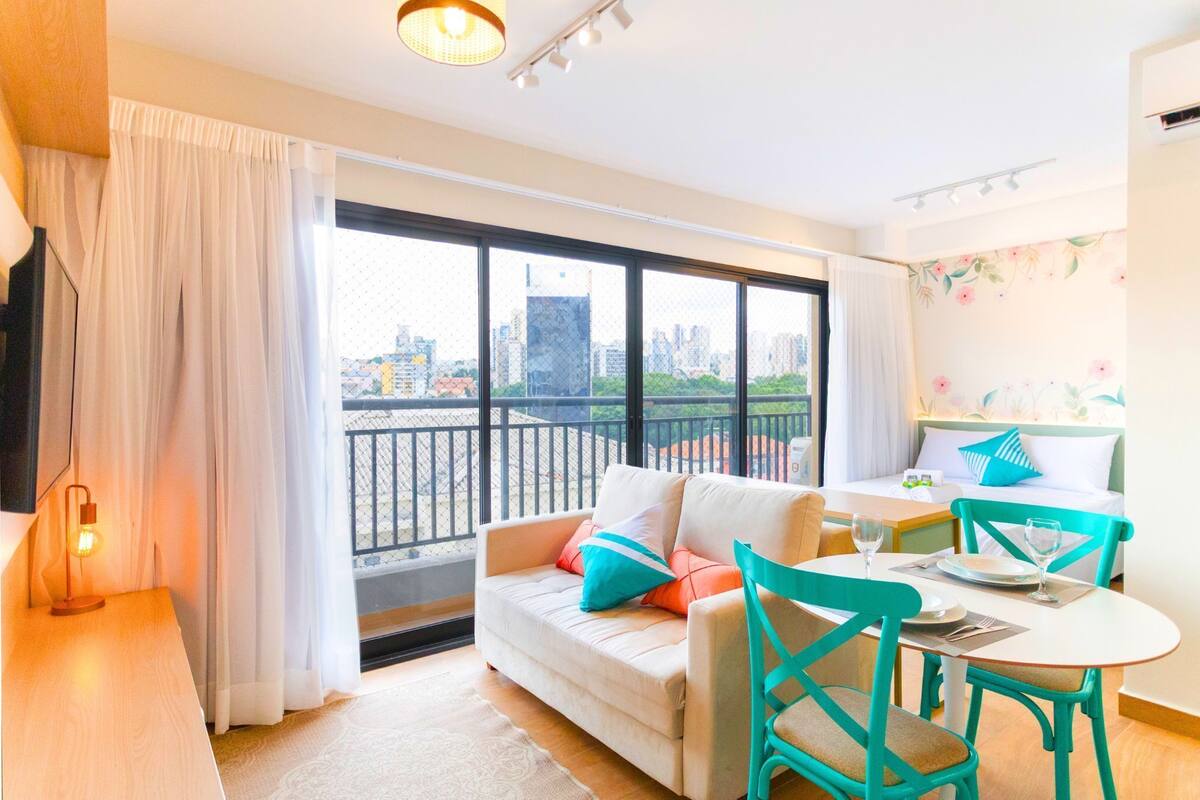 A modern hotel room with a balcony, a sofa, a dining table with chairs, and a city view.