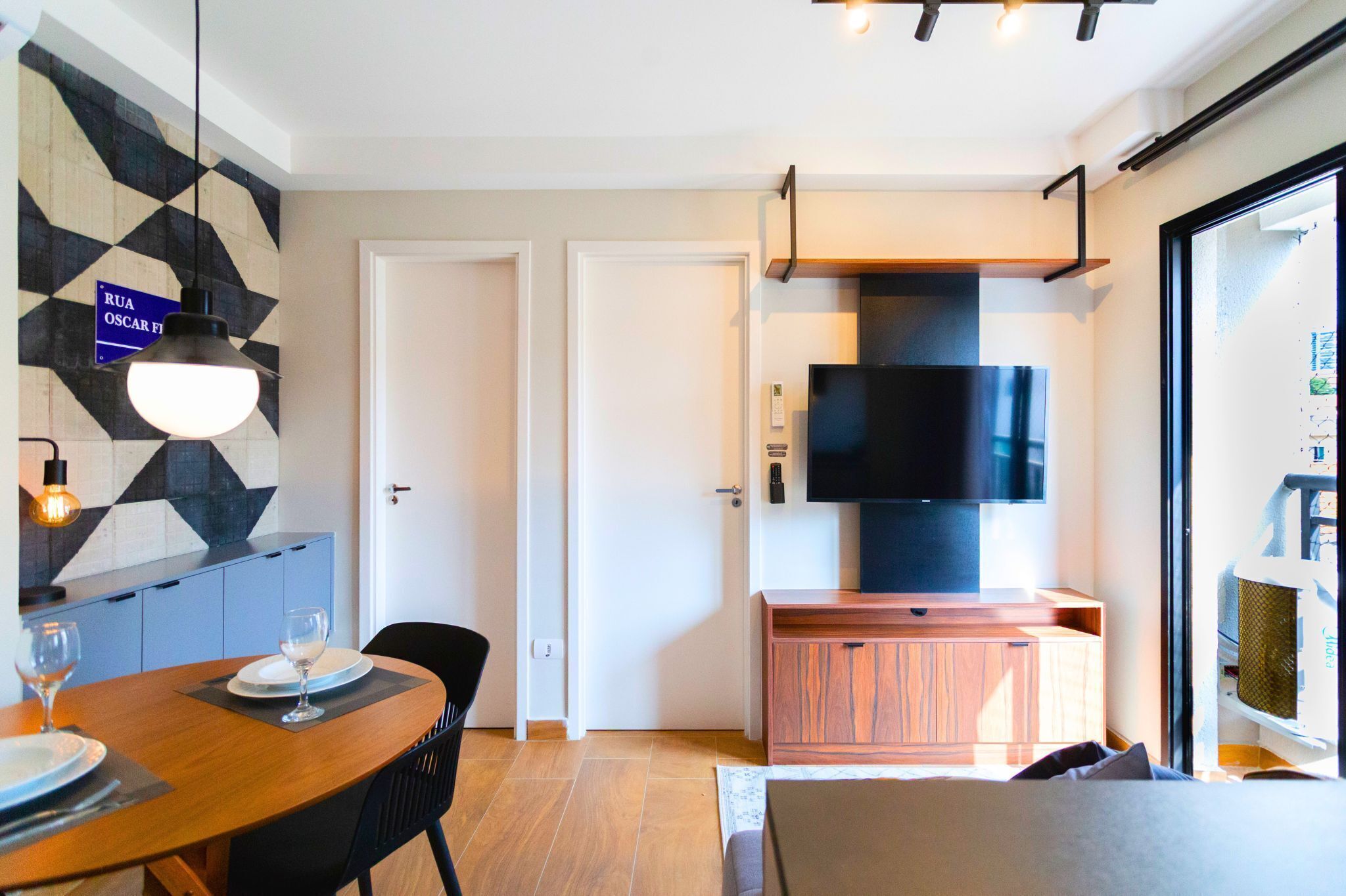 Una habitación de hotel moderna con zona de comedor, un televisor montado en un mueble de madera y una pared con estampado geométrico.