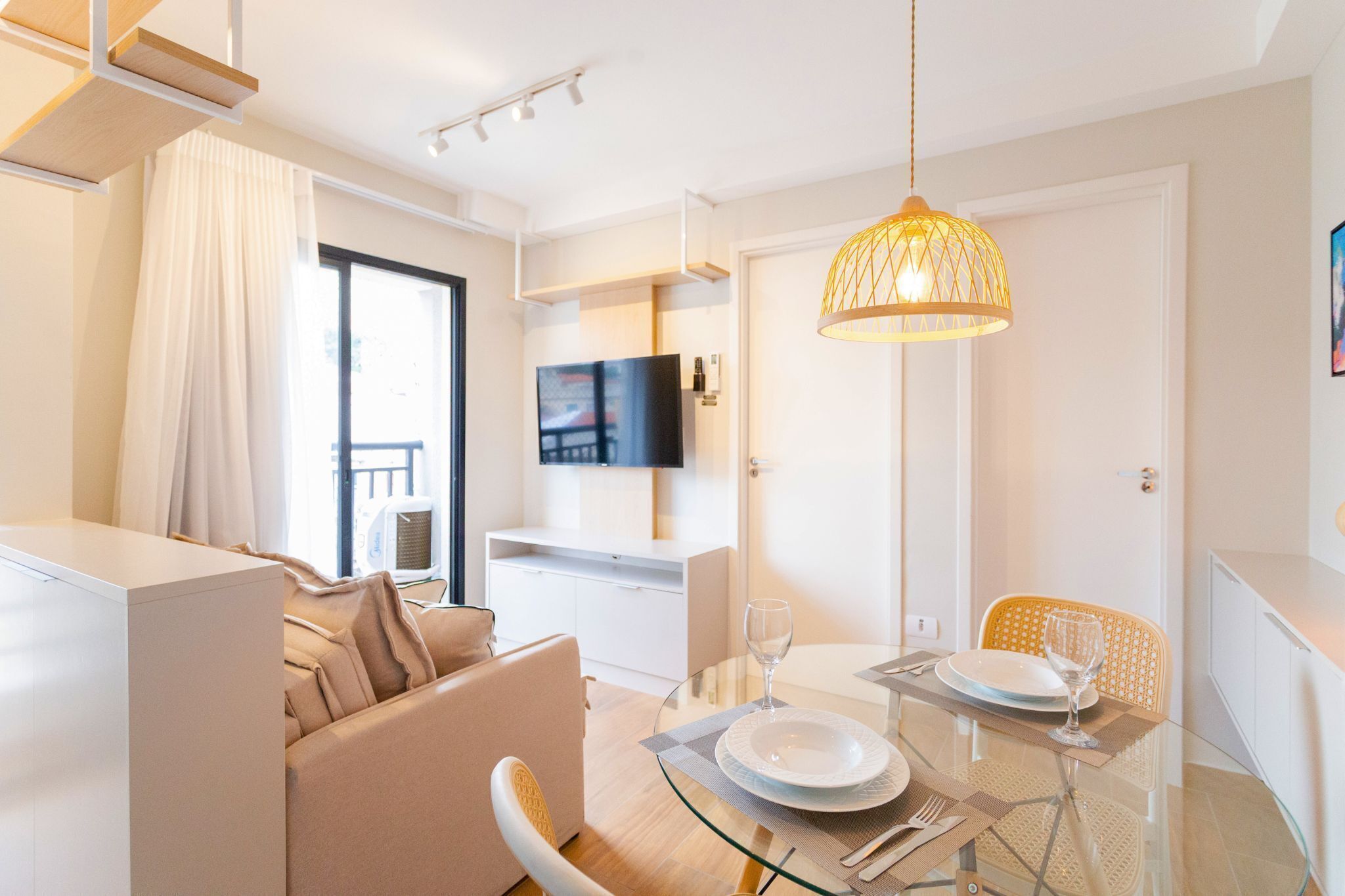 A modern living room with a glass dining table, beige sofa, and a mounted flat-screen TV.