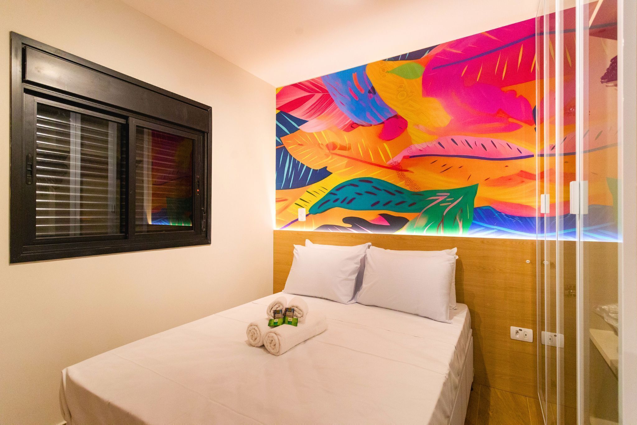 A hotel room with a bed, a window with blinds, and a colorful abstract painting on the wall.