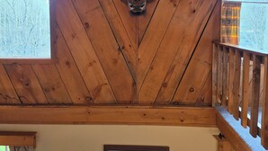 Une pièce avec des poutres apparentes en bois au plafond, une tête d’animal fixée au mur, un tableau encadré et une fenêtre avec des rideaux.