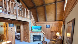 Un intérieur de chalet chaleureux avec une chambre en mezzanine, une cheminée, un téléviseur et un canapé.