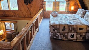 Une chambre mansardée chaleureuse, avec un plafond en pente, un lit recouvert d’une courtepointe matelassée, une décoration murale représentant une tête de taureau et une rambarde en bois.