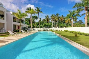 Une piscine entourée de chaises longues, de palmiers et d’un bâtiment blanc.