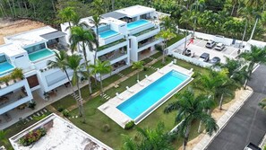 Un immeuble moderne à plusieurs étages, doté d’une grande piscine centrale, entouré de palmiers et d’une pelouse bien entretenue.