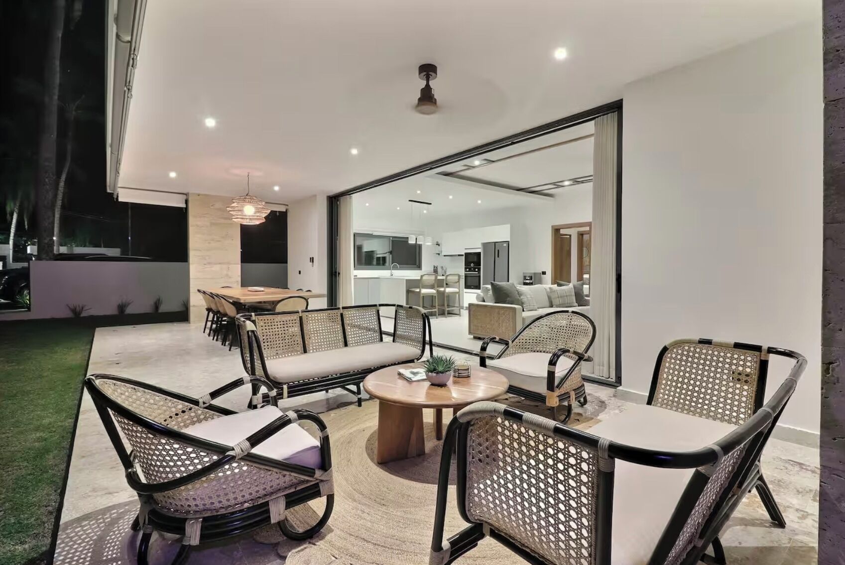 A modern outdoor patio with wicker furniture, a round coffee table, and a glass door leading to a kitchen and living area.