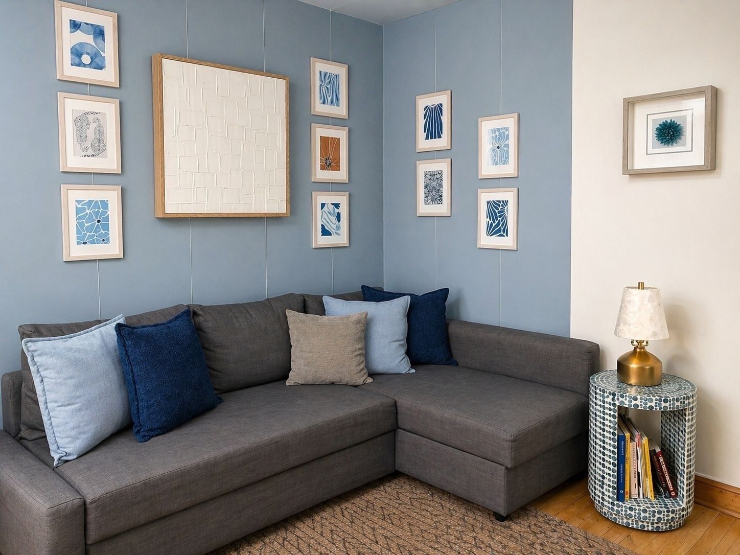 A grey sectional sofa with blue and beige cushions, a patterned rug, and a round side table with a lamp and books.