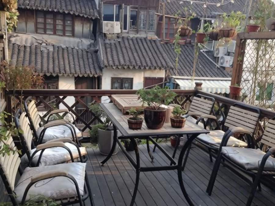 Un balcón con una mesa y sillas, plantas en macetas y techos de tejas tradicionales al fondo.