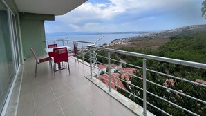 A balcony with a table and chairs, overlooking a coastal town and the sea.