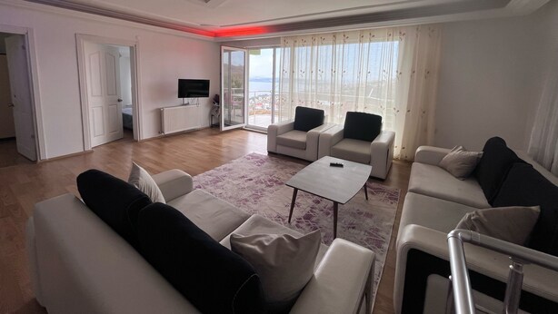 A modern living room with two sofas, a coffee table, and a TV. There is a sliding glass door leading to a balcony with a view.