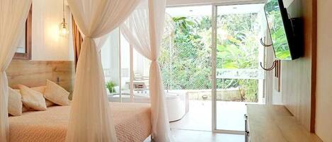 A bedroom with a canopy bed, a flat-screen TV mounted on the wall, and a view of a lush garden through large sliding glass doors.