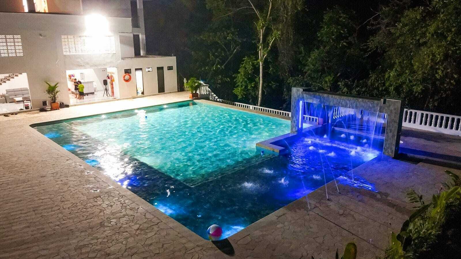A rectangular swimming pool with a waterfall feature, surrounded by a paved deck and a white fence.