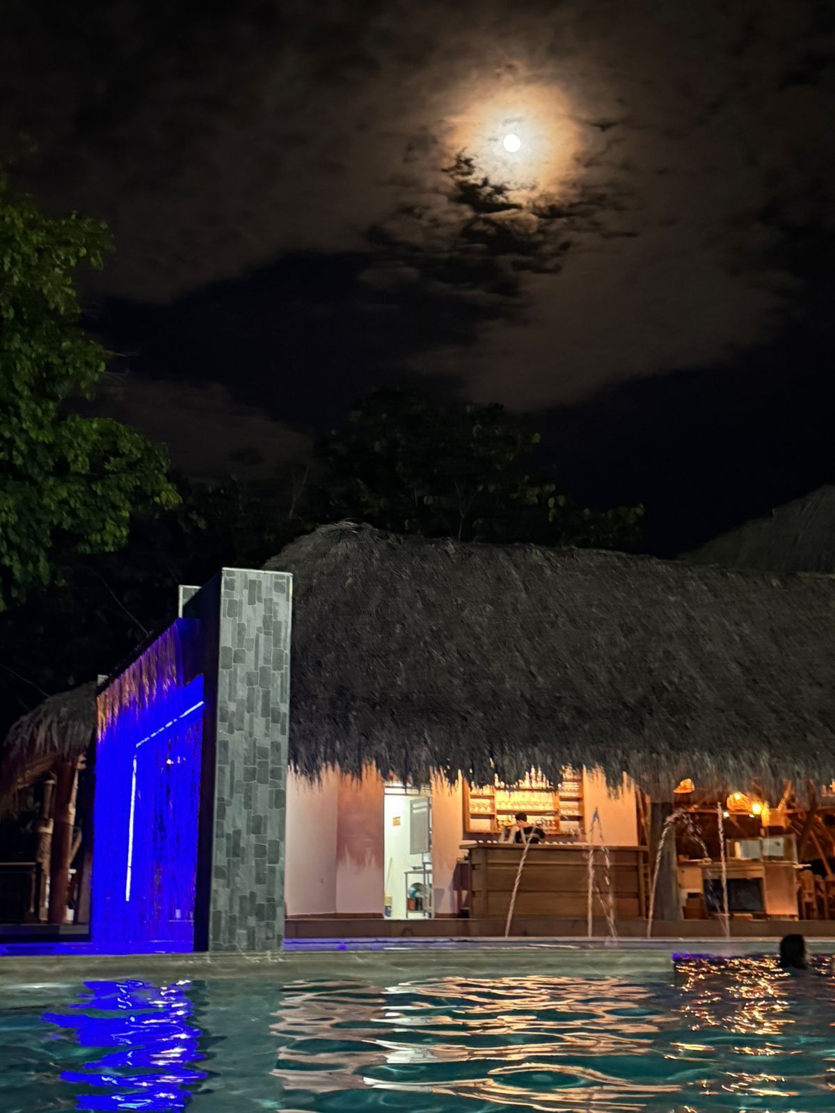 Un área de piscina con una estructura de techo de paja, un pilar de piedra y un cielo iluminado por la luna.