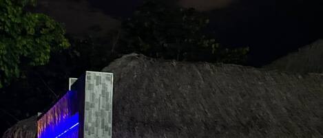 A pool area with a thatched-roof structure, a stone pillar, and a moonlit sky.