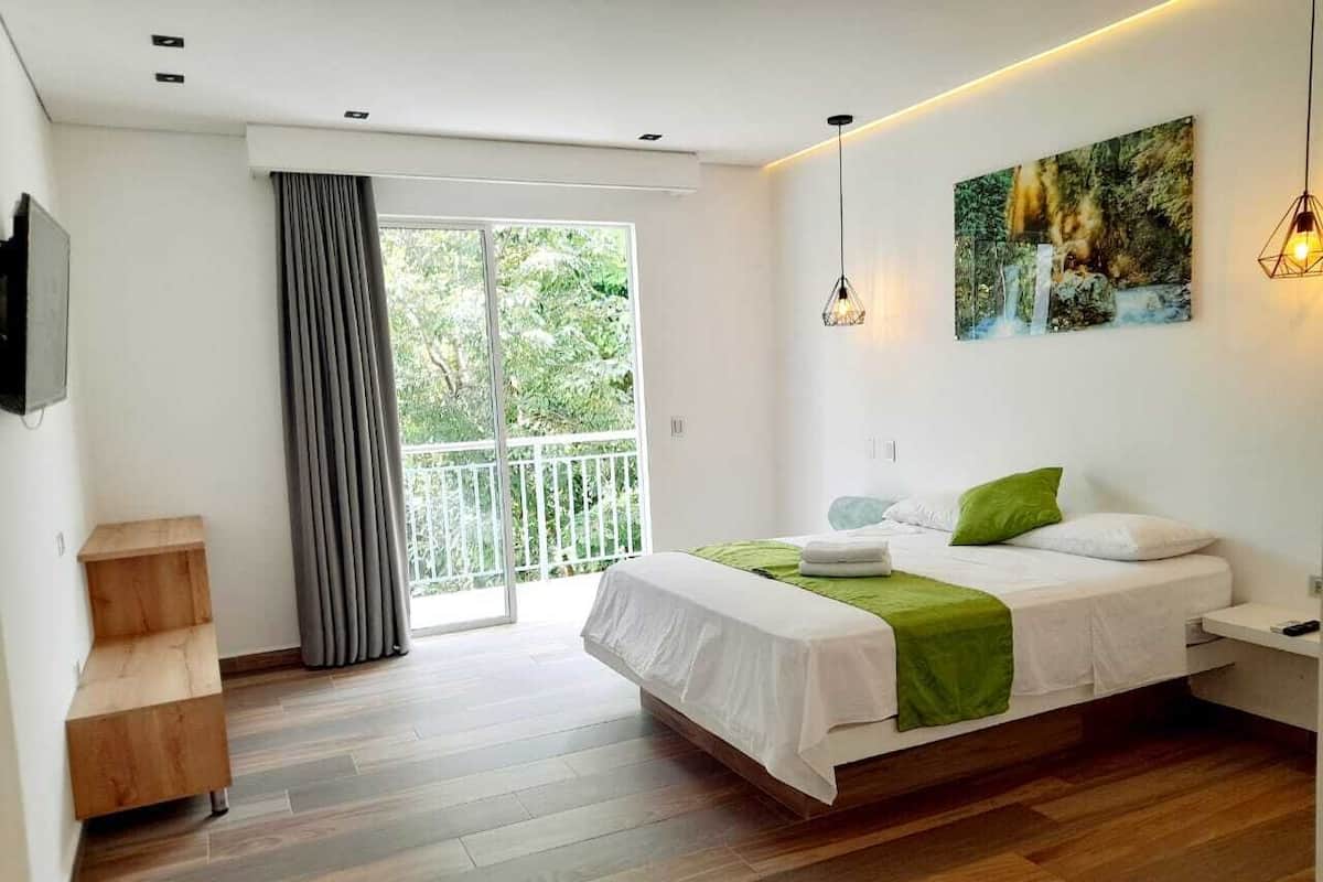 A modern bedroom with a large bed, a wooden bench, a balcony with curtains, and a painting on the wall.