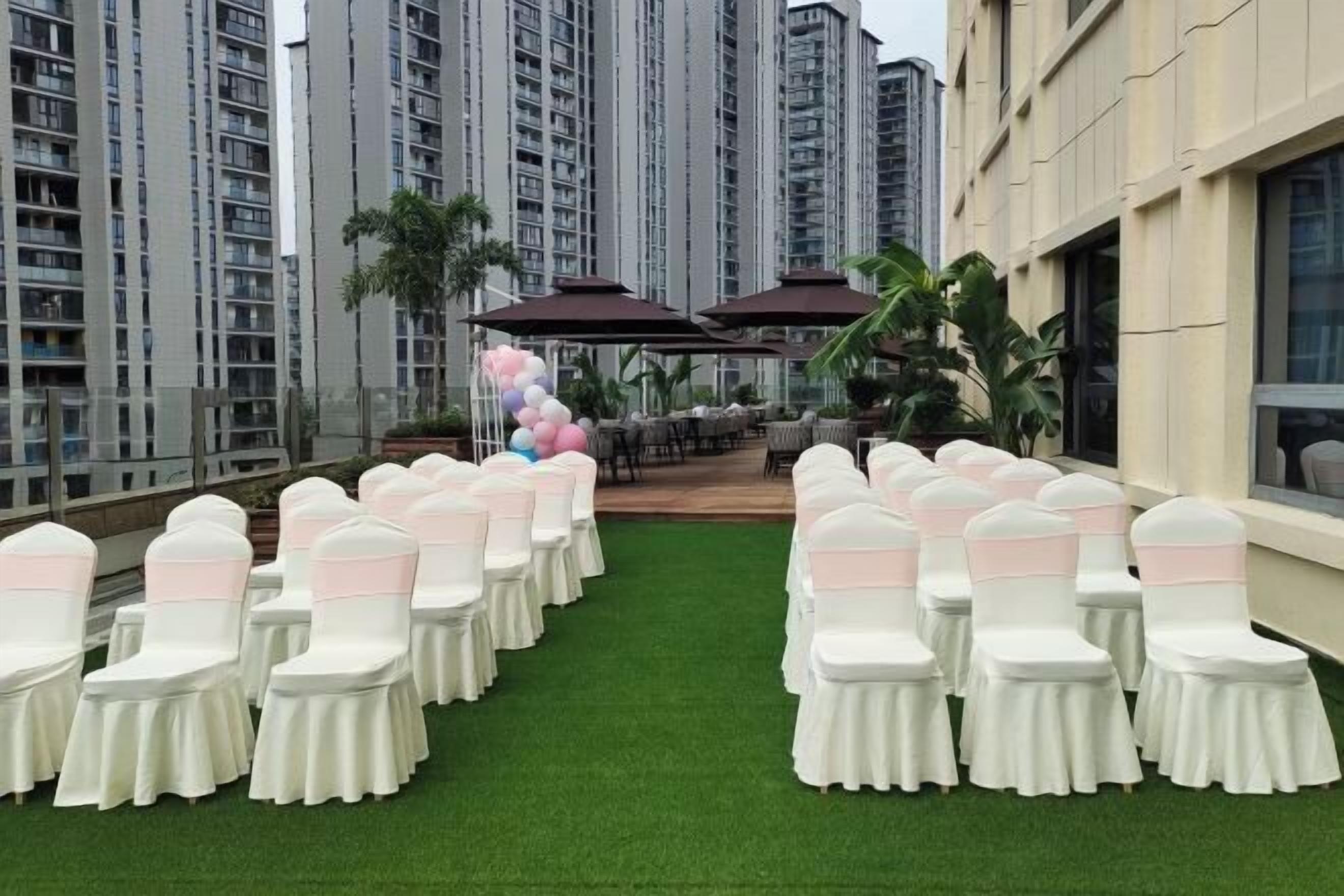 A rooftop venue with rows of white chairs with pink sashes, a green artificial turf, and a backdrop of tall residential buildings.