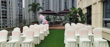 A rooftop venue with rows of white chairs with pink sashes, a green artificial turf, and a backdrop of tall residential buildings.