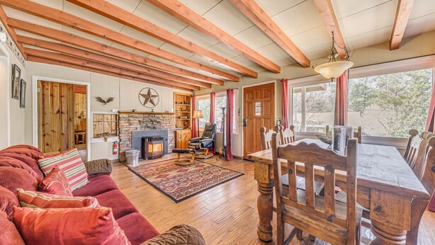 A cozy living room with a fireplace, wooden beams, a dining table with chairs, and a large window. - Charming 2-bedroom house in Ruidoso with AC, WiFI, pellet stove, hot tub (Ruidoso)