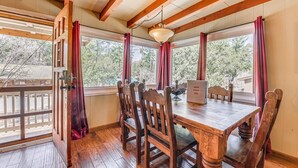 A wooden dining table with six chairs, a chandelier, and large windows with red curtains. - Charming 2-bedroom house in Ruidoso with AC, WiFI, pellet stove, hot tub (Ruidoso)