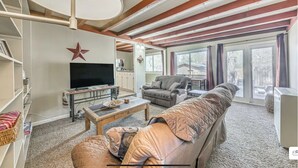 A spacious living room with a large flat-screen TV, a sofa, and a coffee table. There is a star decoration on the wall and a window with curtains. - Charming 2-bedroom house in Ruidoso with AC, WiFI, pellet stove, hot tub (Ruidoso)