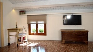 Una habitación con un escritorio de madera, una silla, un banco, un televisor montado en la pared y una ventana con vistas a zonas verdes.