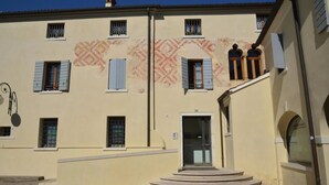Un edificio con fachada color crema, varias ventanas con contraventanas, una entrada central con escalones y un pequeño balcón con dos personas.