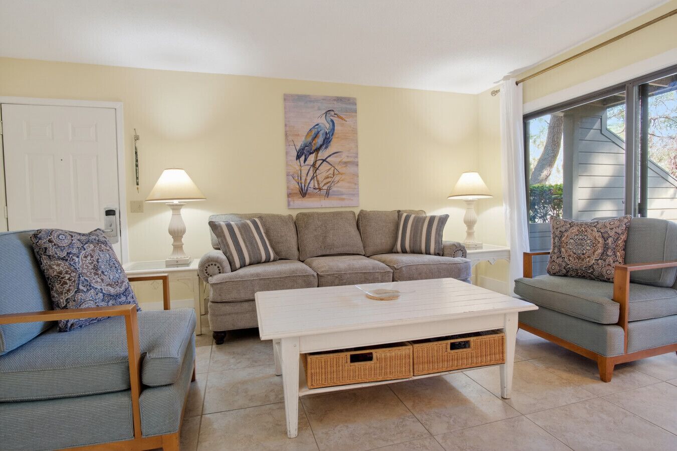 Un salon avec un canapé gris, une table basse blanche et deux fauteuils. Un tableau représentant un oiseau est accroché au mur, et une porte est visible au fond.