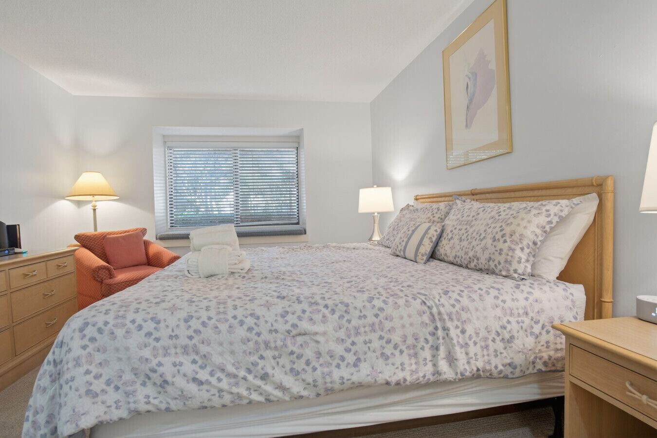 Une chambre à coucher avec un lit, deux lampes de chevet, une fenêtre avec des stores, une chaise, un bureau et un tableau encadré au mur.