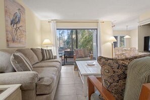 Un salon comprenant un canapé, un fauteuil à motifs, une table basse et un coin repas avec des chaises et une table. Un tableau orne le mur et, à travers les portes vitrées, on peut admirer la vue sur l’extérieur.