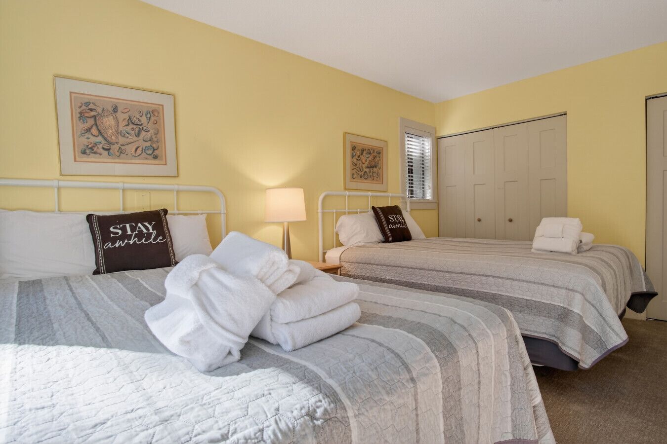 A hotel room with two beds, each with a striped bedspread and pillows with text. There are two framed pictures on the walls, a window with blinds, and a door.