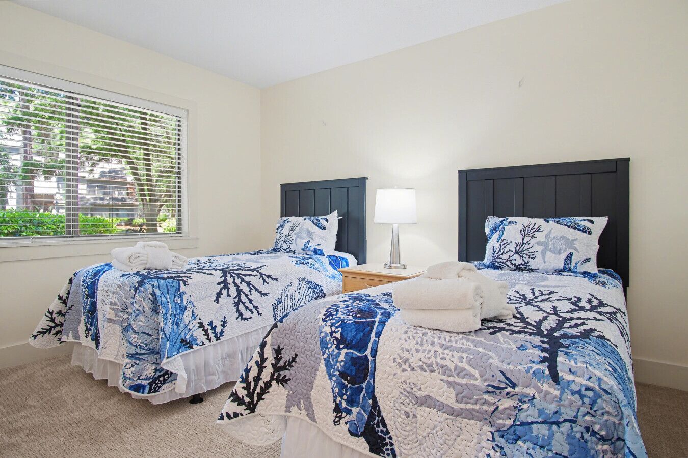 A hotel room with two beds, a window with blinds, a bedside lamp, and a view of greenery outside.