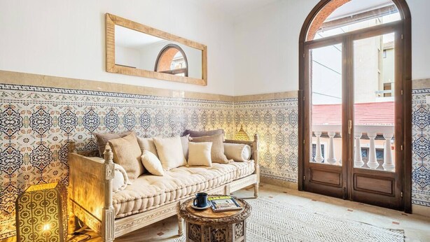 Ein Zimmer mit einer Wand aus gemusterten Fliesen, einem beigen Sofa mit Kissen, einem kleinen Holztisch und einem großen, bogenförmigen Durchgang.