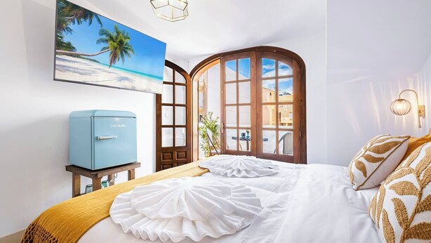 Een slaapkamer met een bed, een blauwe minibar, een schilderij van een strand en een raam met uitzicht op gebouwen.