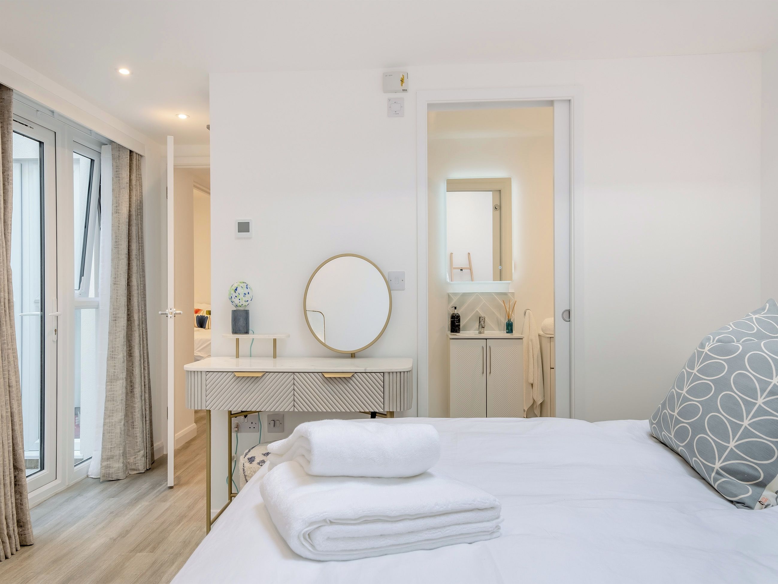 Ein Schlafzimmer mit Bett, Nachttisch, rundem Spiegel, Fenster mit Vorhängen und angrenzendem Badezimmer.