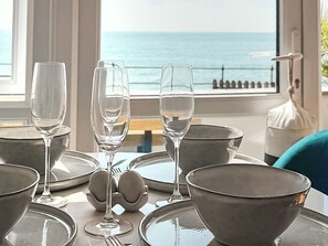 A table set with wine glasses and bowls, a window with a view of the sea, and a white birdcage.