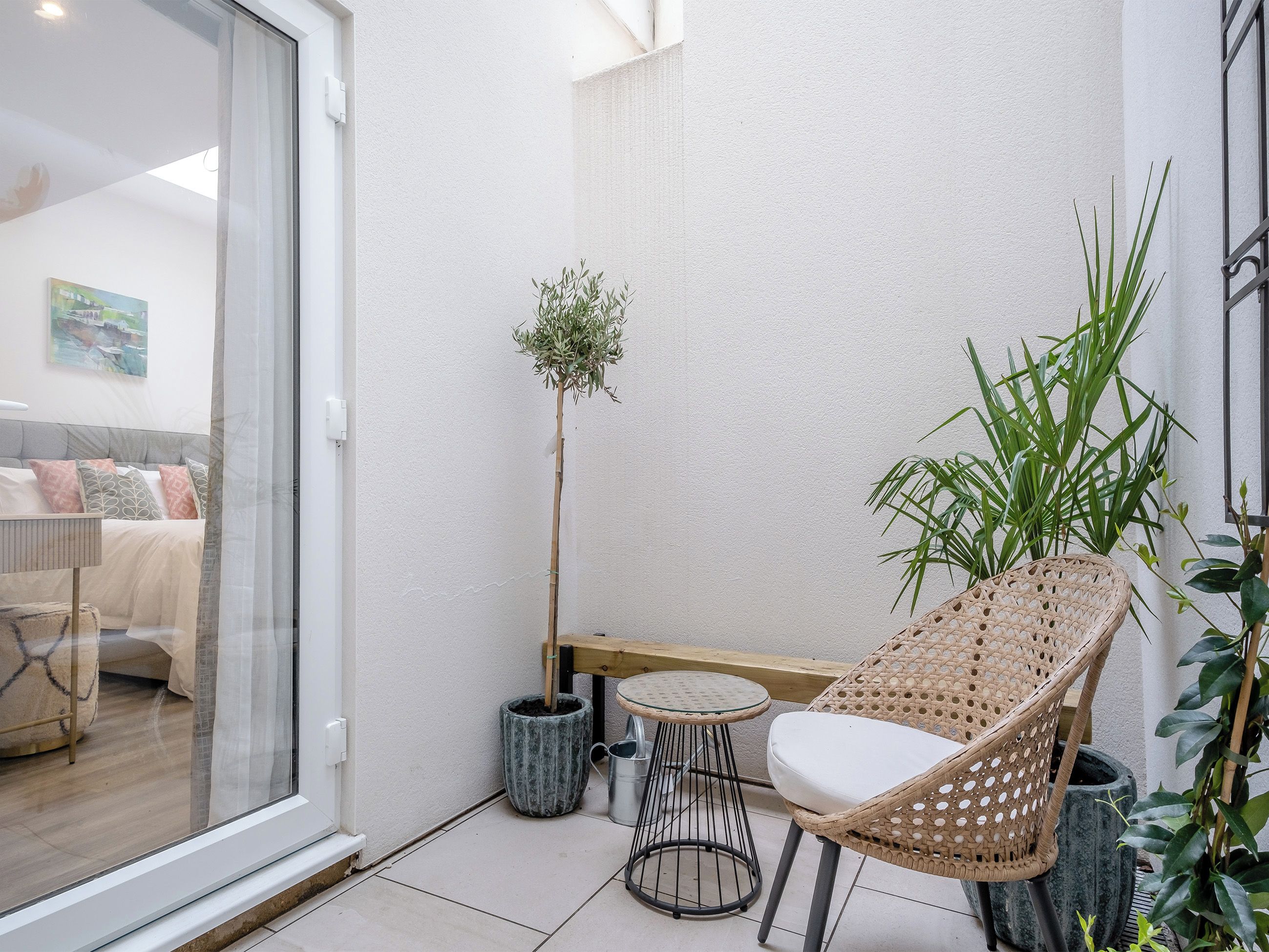 A small balcony with a wicker chair, a potted plant, and a glass door leading to a bedroom.