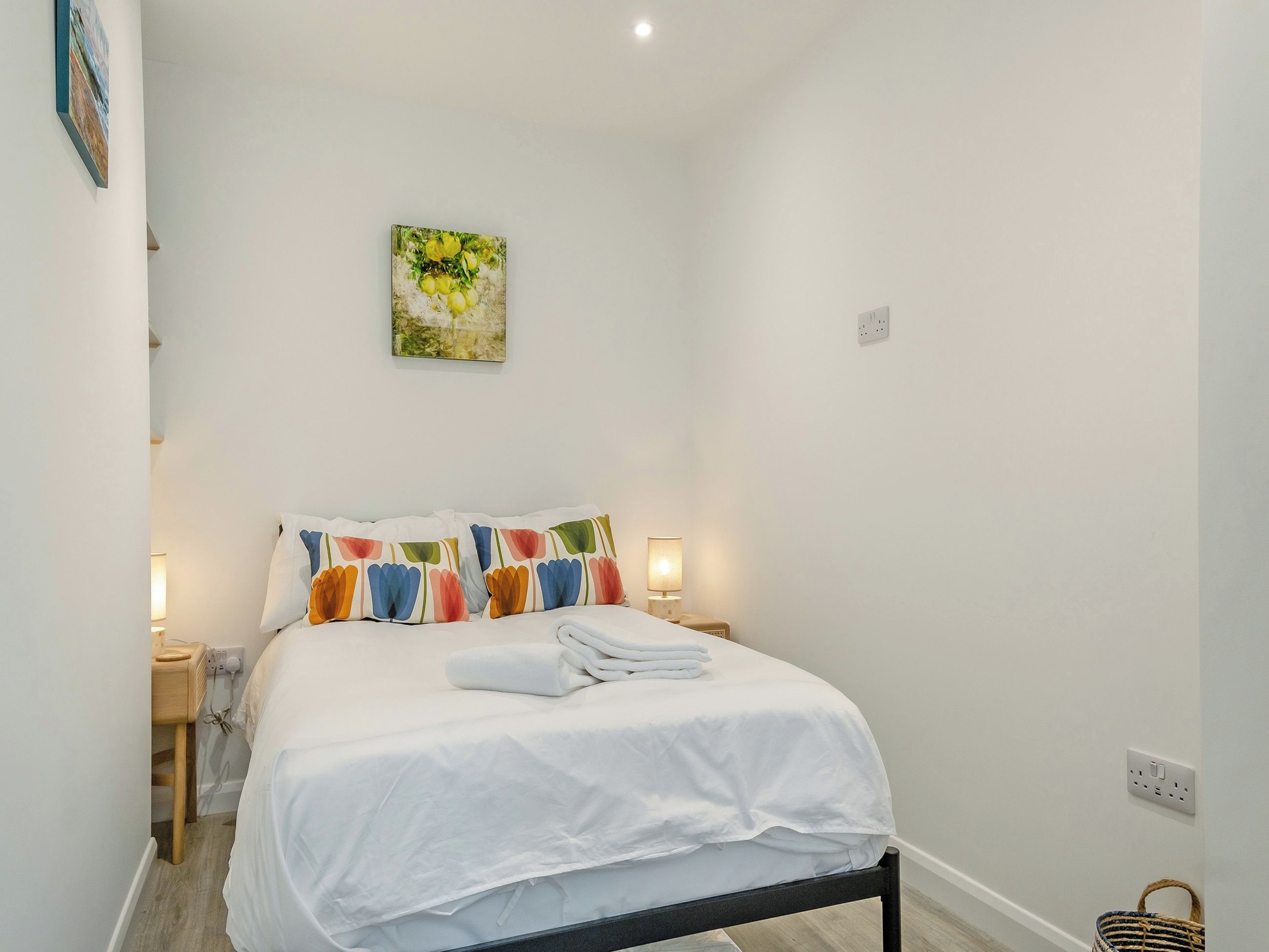 A neatly made bed with white bedding and colorful pillows, a bedside table with a lamp, and a painting on the wall.