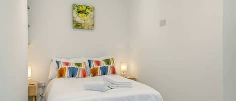Una cama bien hecha con ropa blanca y almohadas coloridas, una mesita de noche con una lámpara, y un cuadro en la pared.