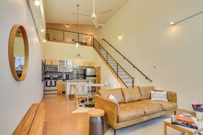Un vaste espace de vie comprenant un canapé en cuir, une table basse et une table à manger avec des chaises. On aperçoit une cuisine au fond, et un escalier mène à l’étage supérieur.