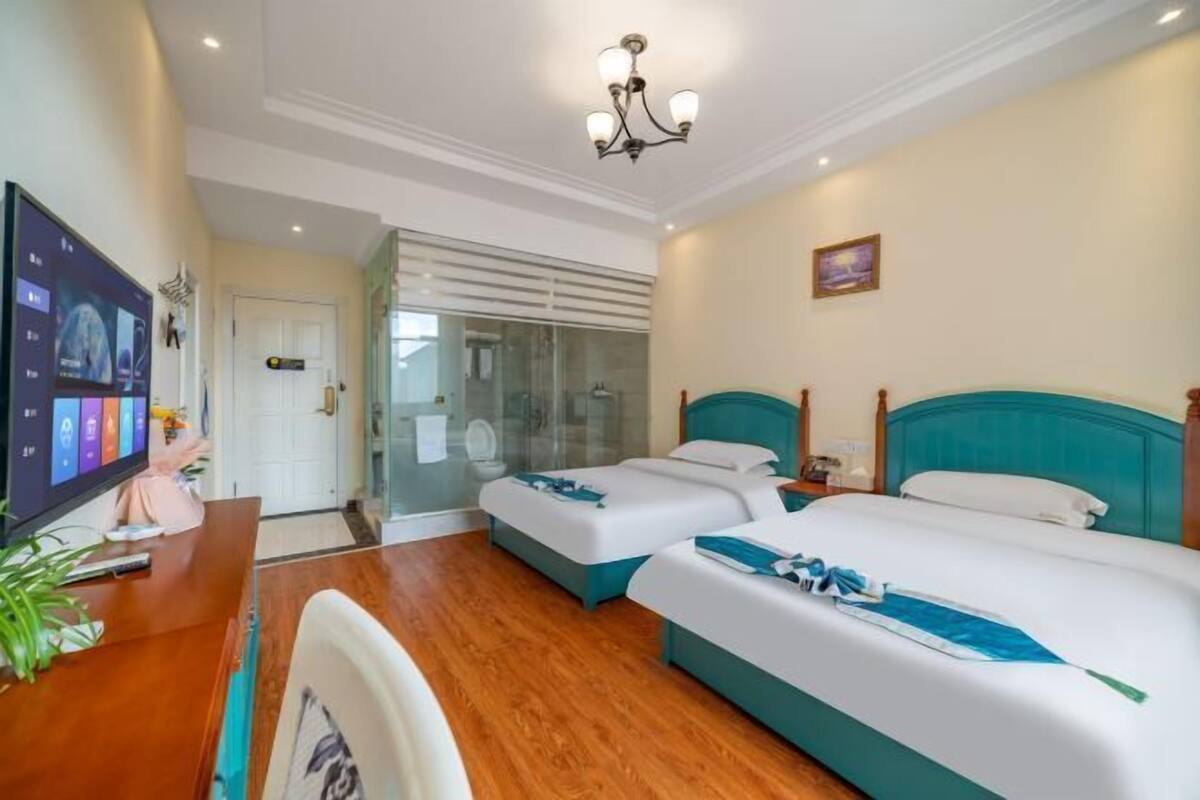 Ein Hotelzimmer mit zwei Betten, einem Schreibtisch mit Computer, einem Fernseher und einem Badezimmer mit Glastür.