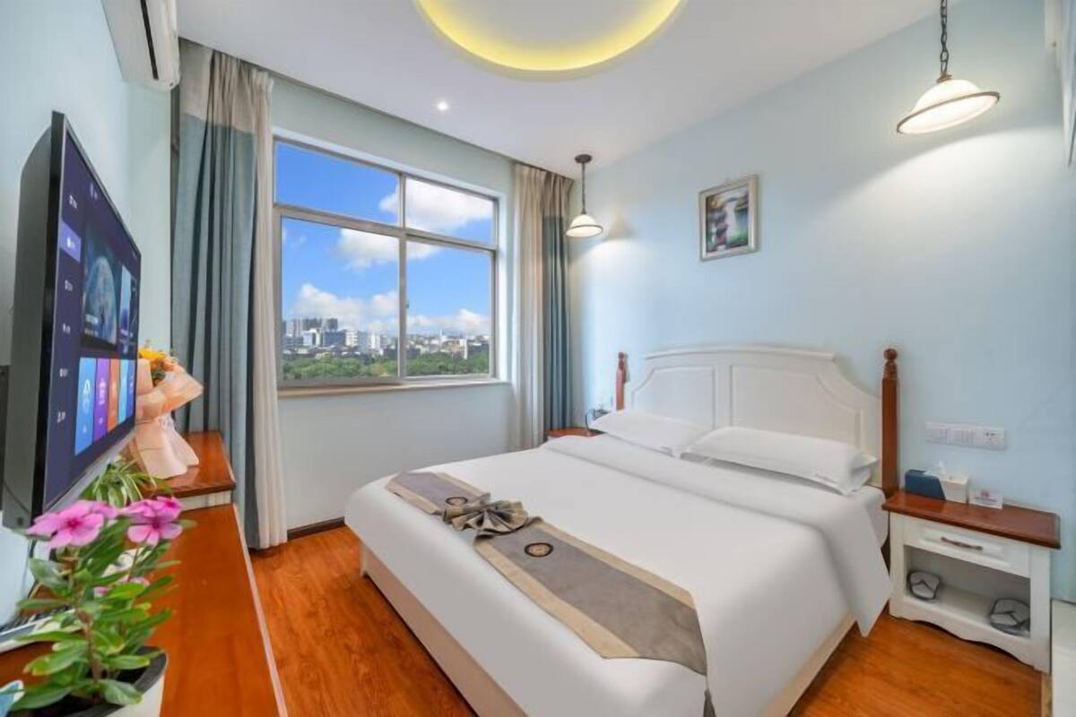 Ein Hotelzimmer mit einem großen Bett, einem Fernseher, einem Fenster mit Blick auf die Stadt und einem Nachttisch mit Lampe.