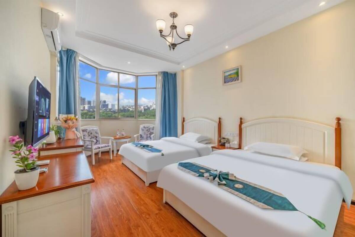 Ein Hotelzimmer mit zwei Betten, einem Schreibtisch mit Fernseher, einem Sessel, einem kleinen Tisch und Blick auf die Stadt.