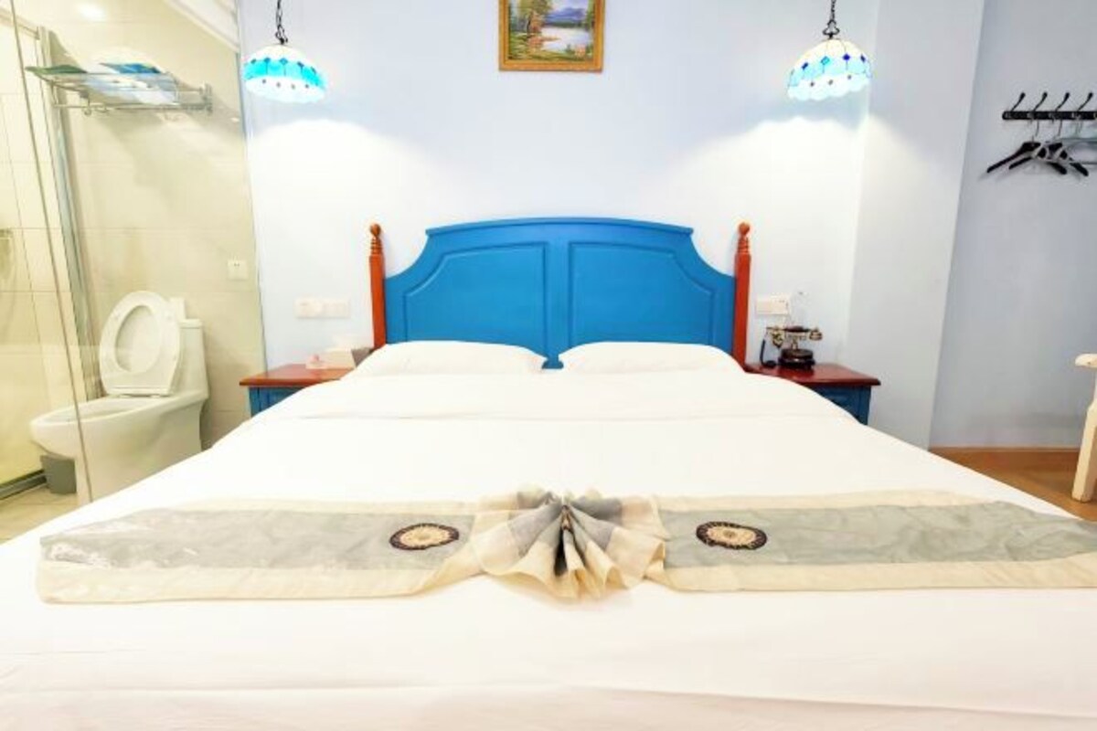 A hotel room with a large bed, a wooden headboard, a bedside table with a lamp, and a bathroom with a glass shower enclosure.