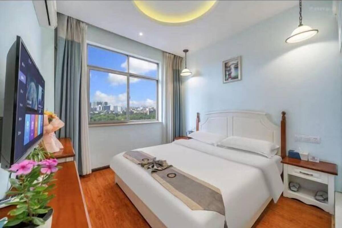 Ein Hotelzimmer mit großem Fenster, einem Bett, einem Nachttisch, einem Fernseher und einer Pflanze.