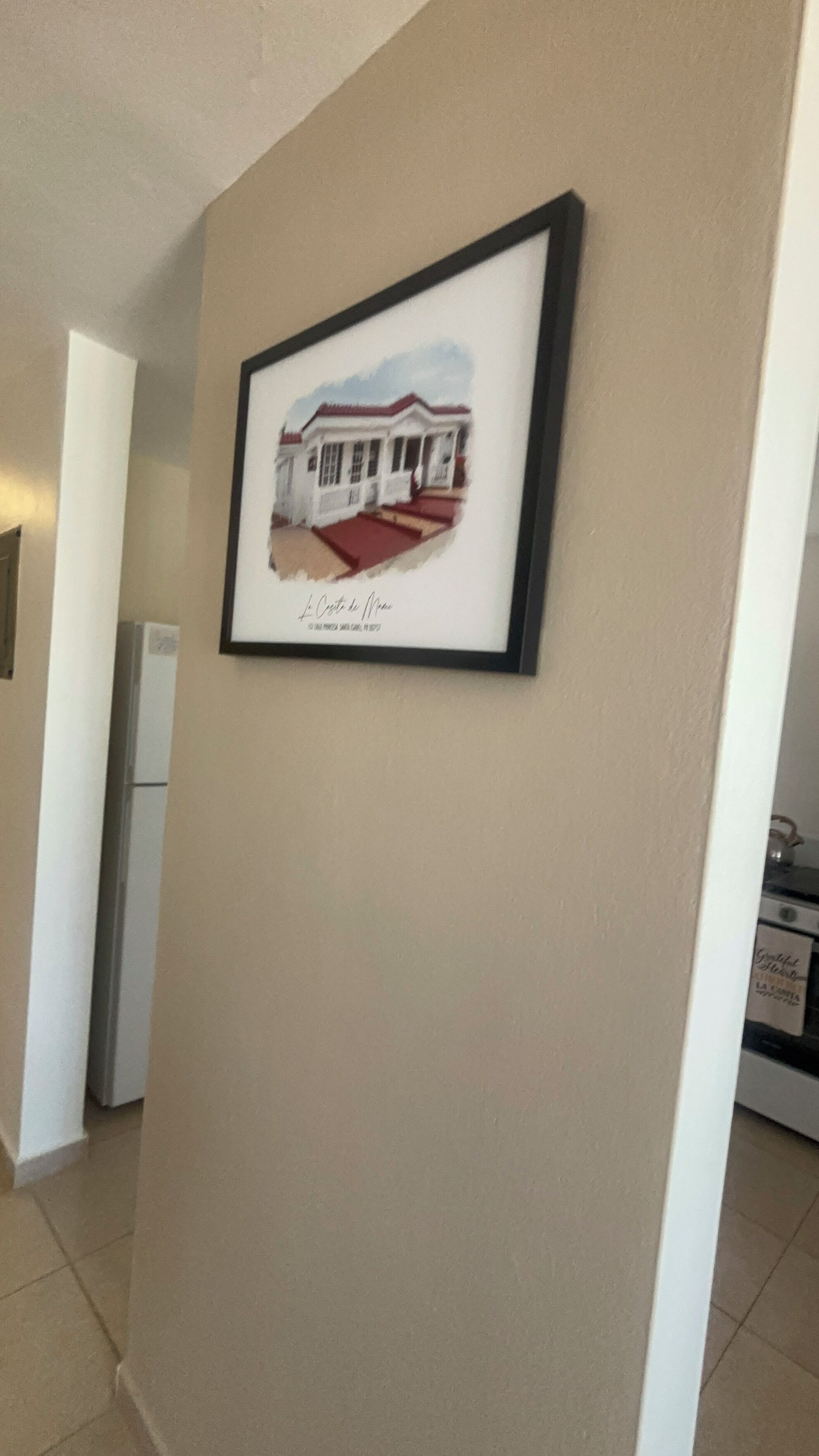 A framed picture of a house on a beige wall in a hallway.