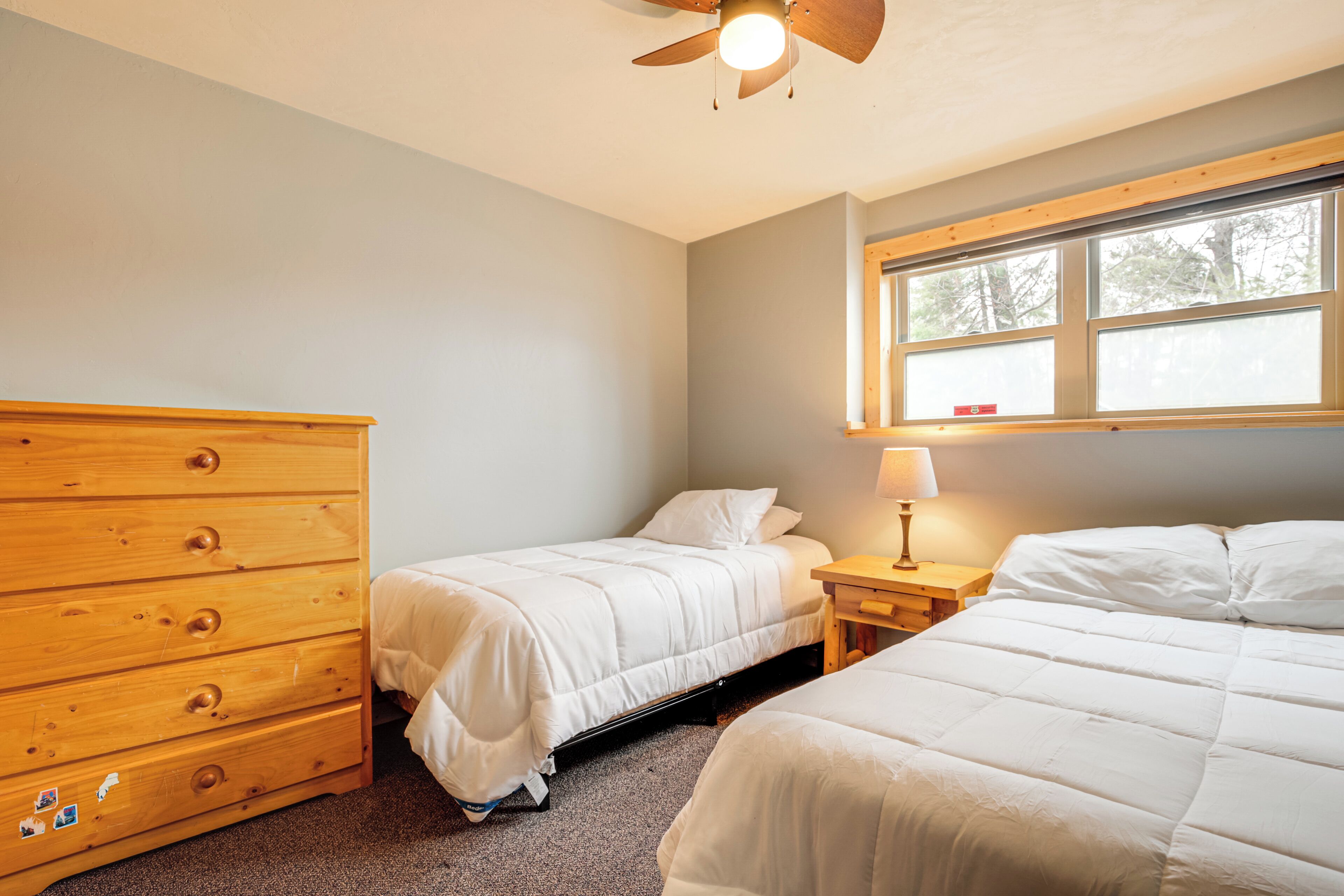 A bedroom with two beds, a wooden dresser, a nightstand with a lamp, and a window with a view of trees.
