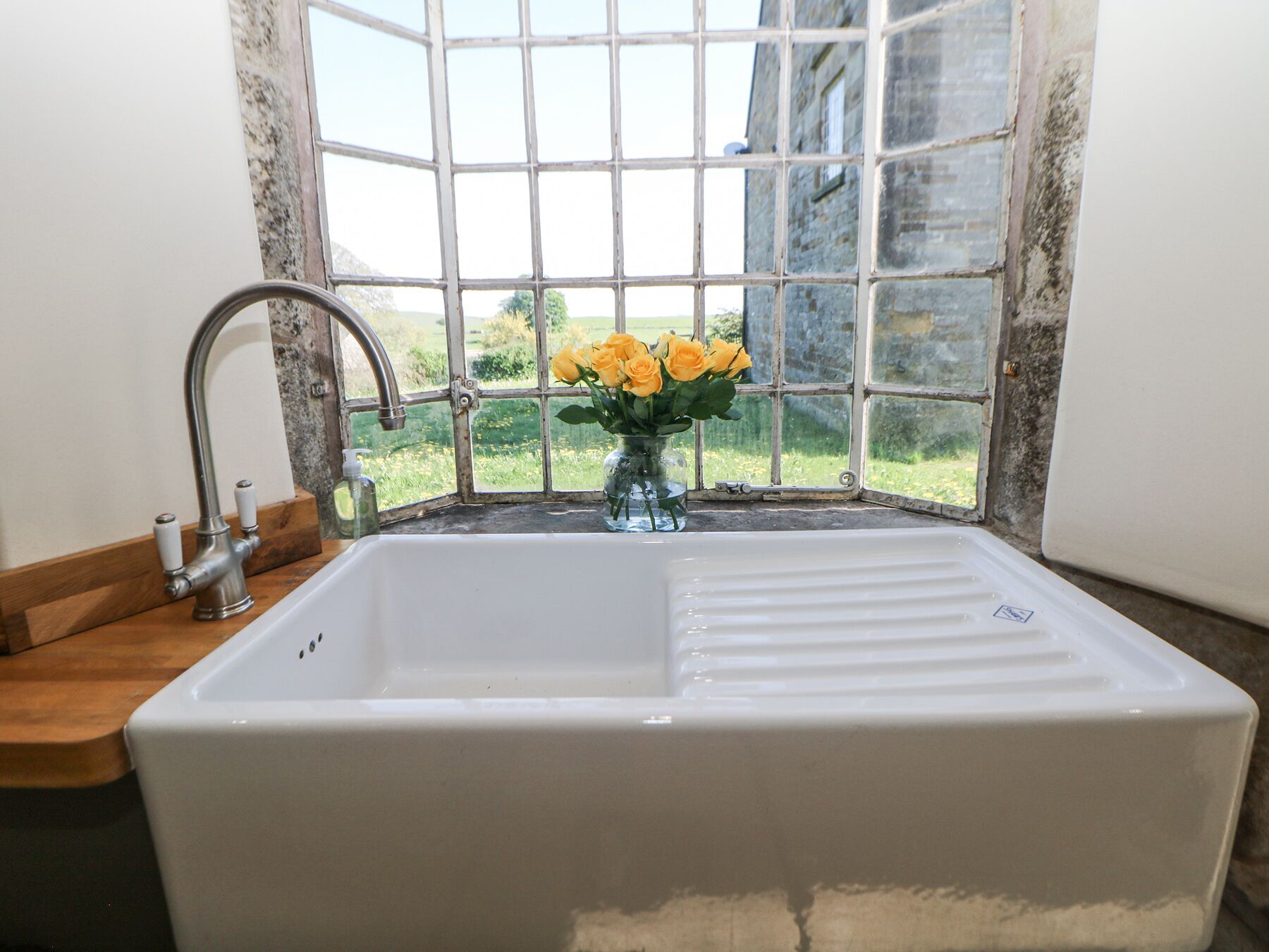 A bathroom with a white sink, a faucet, a glass vase with yellow flowers, and a view of greenery through a window with a grid pattern.