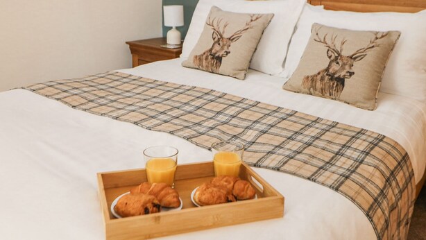 Ein ordentlich bezogenes Bett mit einer Karo-Decke, zwei Kissen mit Hirschmotiven, ein Nachttisch mit Lampe und eine Ablage mit Croissants und Saft.