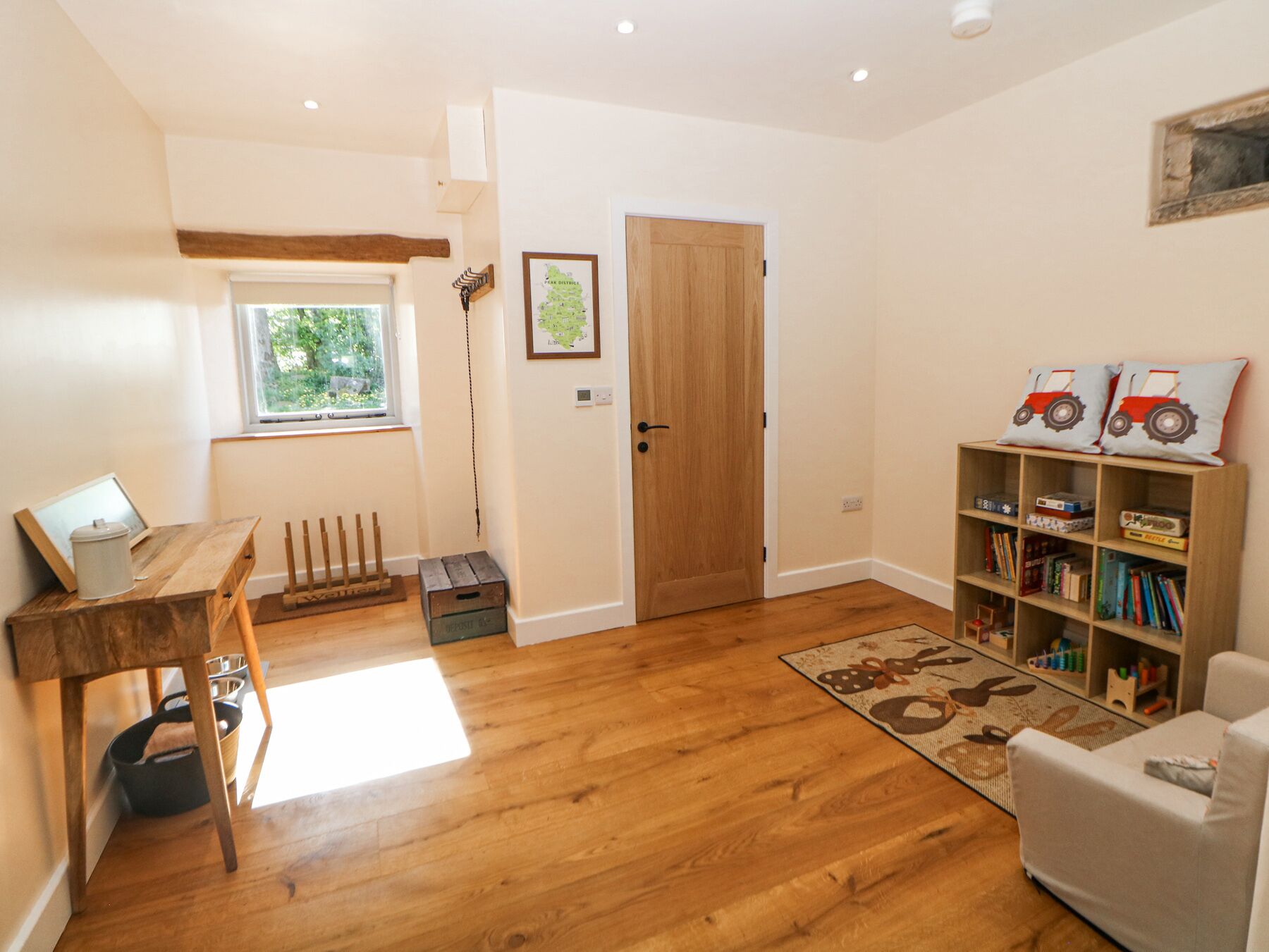 A cozy room with wooden flooring, a wooden desk, a bookshelf with children's toys, a door, and a window with a view of greenery.